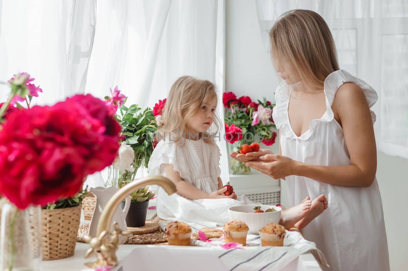 A little blonde girl with her mom on a kitchen countertop decorated with peonies. The concept of the relationship between mother and daughter. Spring atmosphere. by Annu1tochka