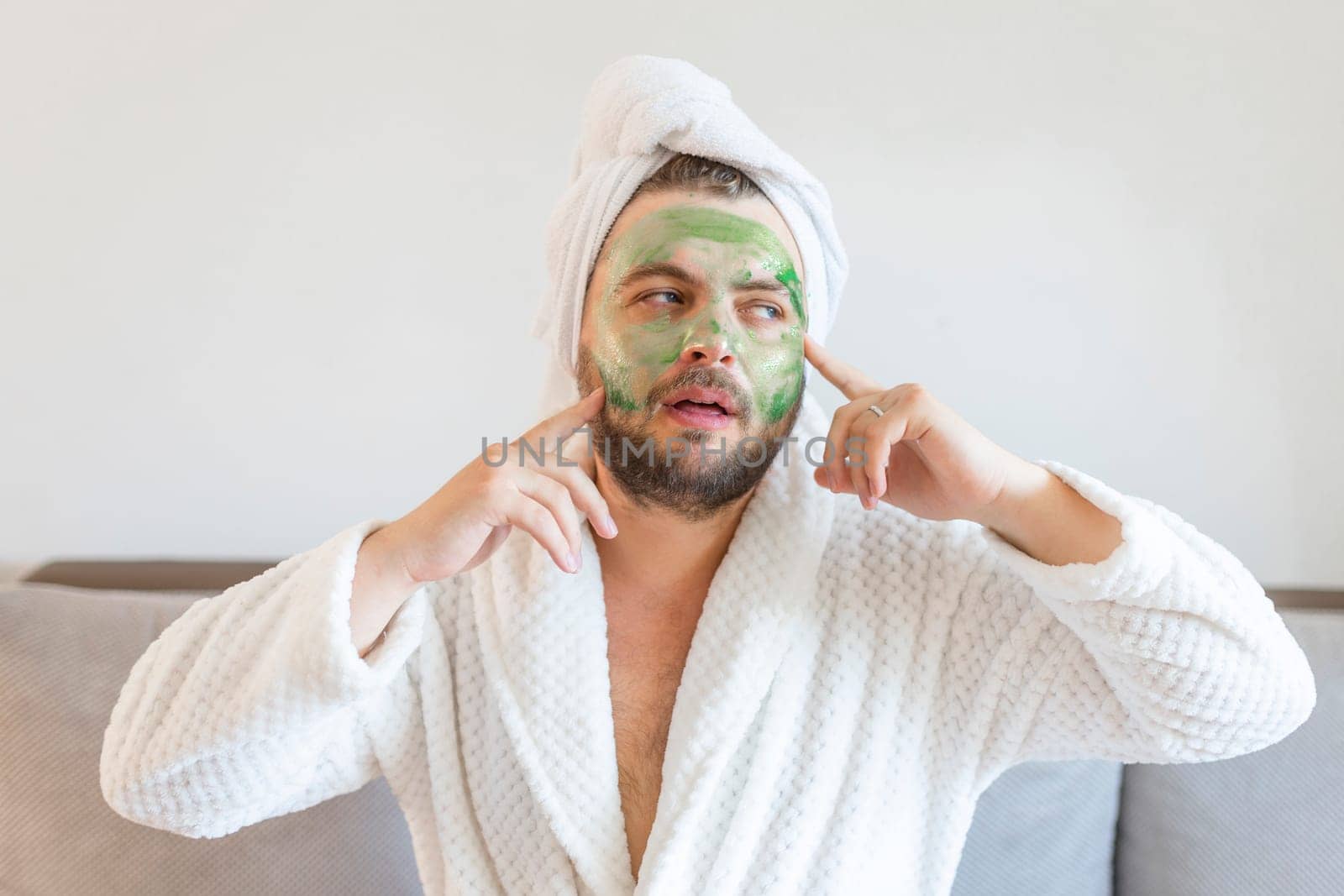 Surprised guy spends hours treating and caring for his skin, applies green face mask to pamper himself, wearing a soft bathrobe and a towel on his head.