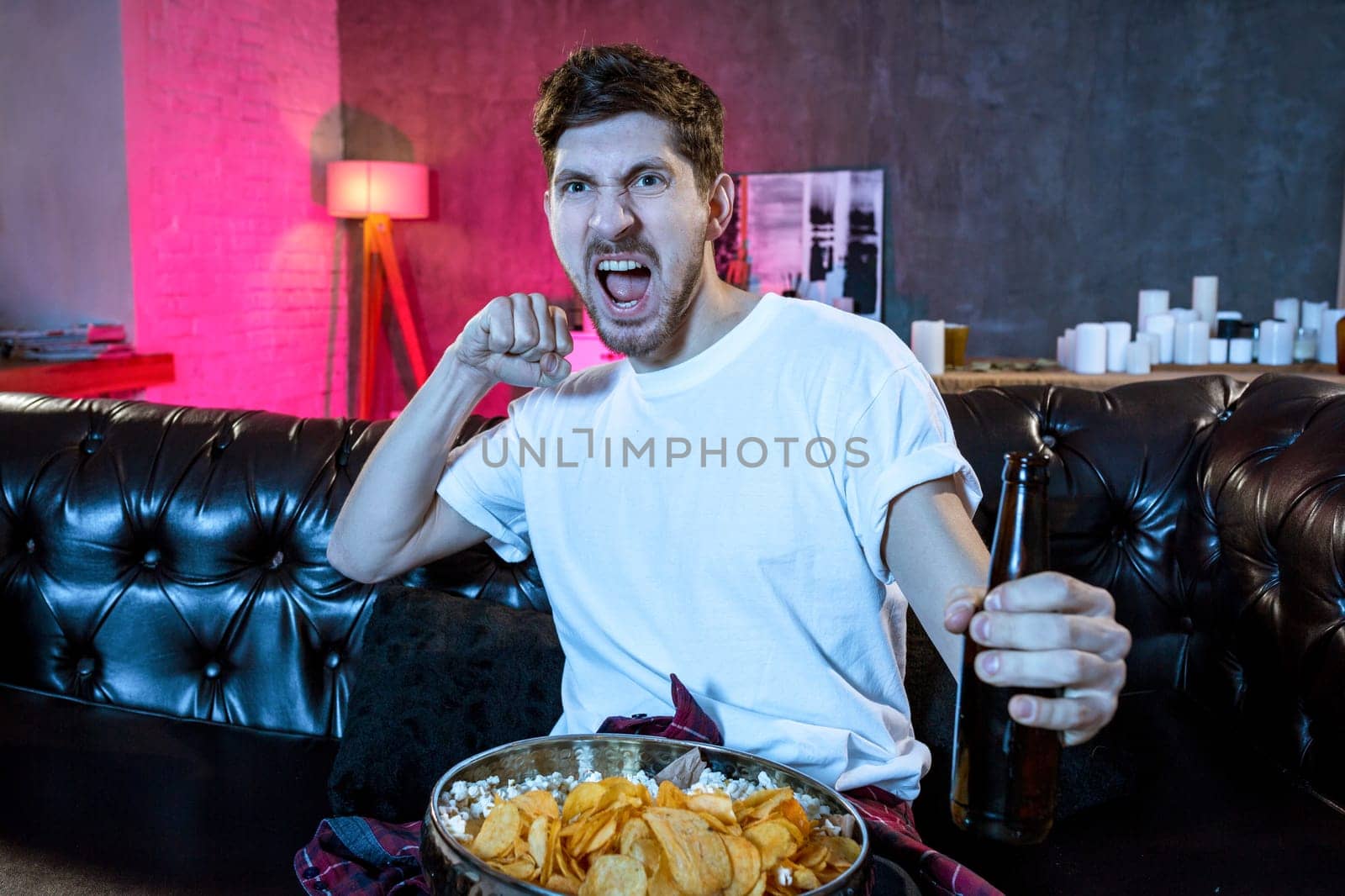Young fan man watching football game on television celebrating goal happy on sofa couch at home with beer bottle in crazy enthusiastic face expression