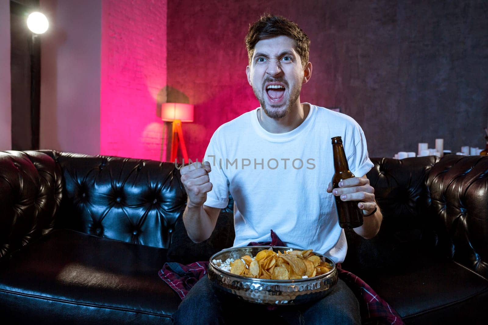 Young supporter man watching football game on television sitting at home couch with beer and chips in stress dejected and disappointed for failure shouting and complaining.