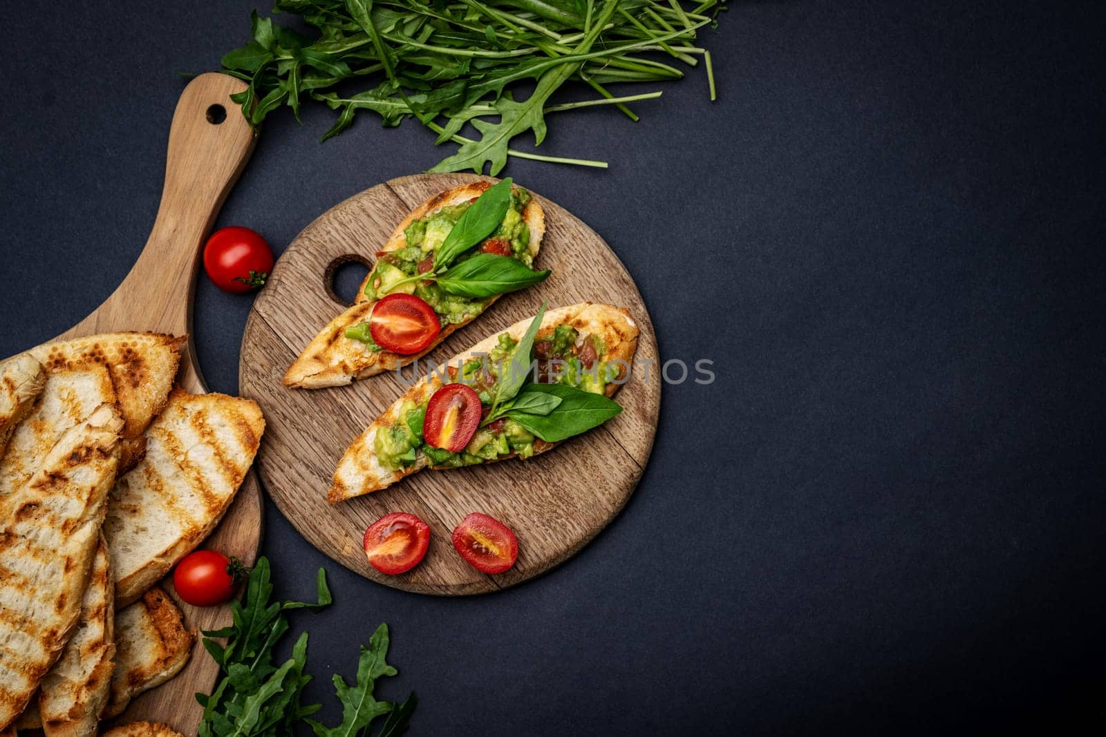 Bruschettas with toast, guacamole, basil and cherry tomatoes on wooden board served with arugula. Mexican snack with french slice bread with avocado, herbs and vegetables on background with copy space