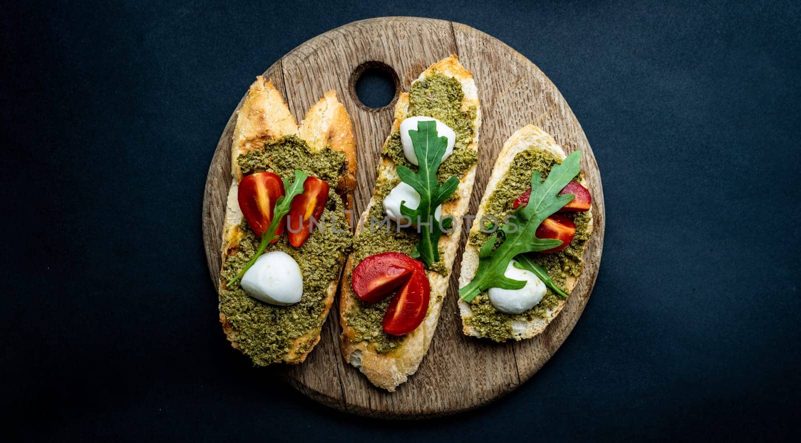 Bruschettas with pesto, tomatoes, mozzarella cheese and basil on wooden board. Italian tosted bread with vegetables and traditional sauce