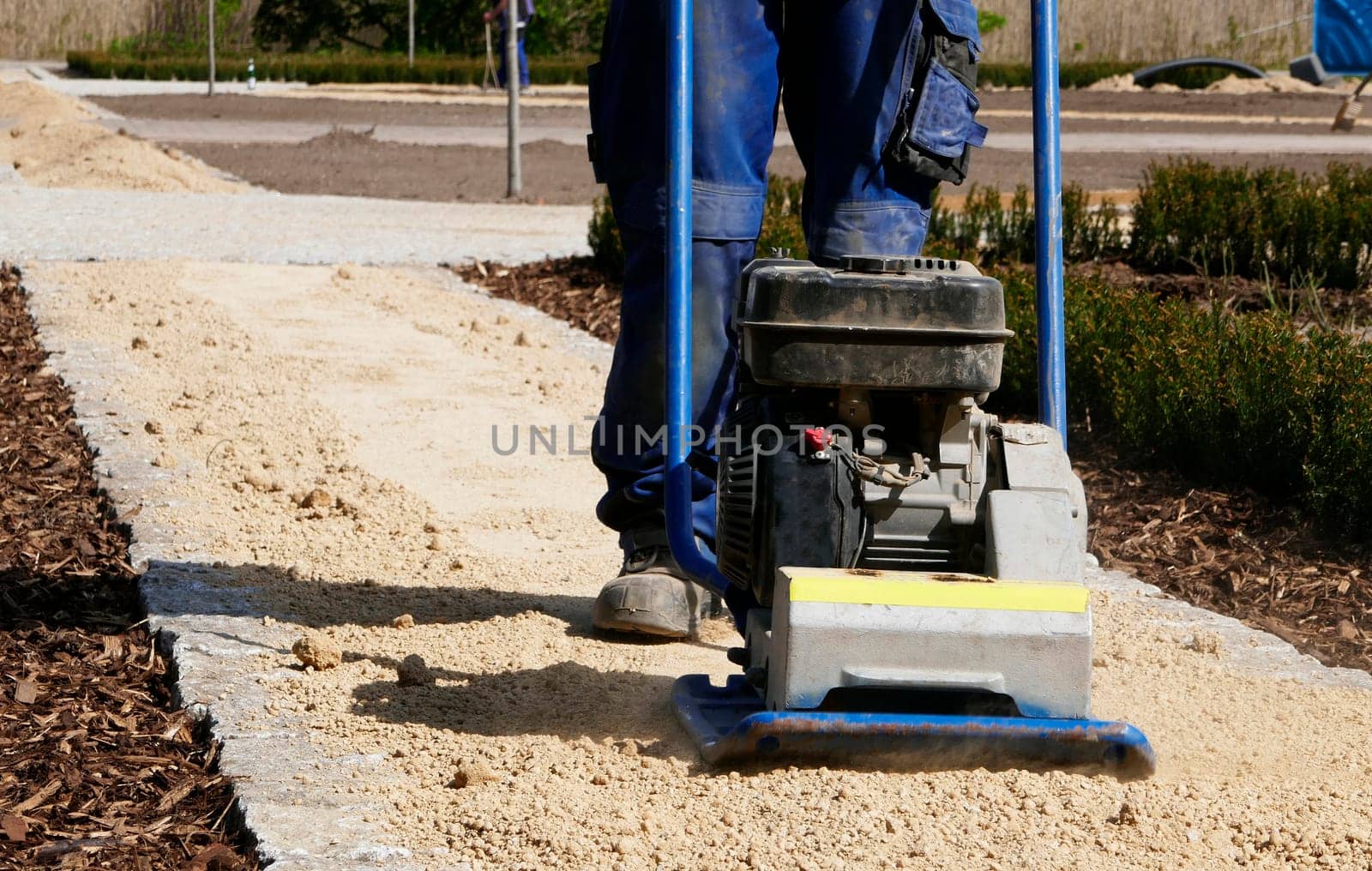 Legs and feet of a man who operates a sand compactor. The man is vibrating the sand of a new path.