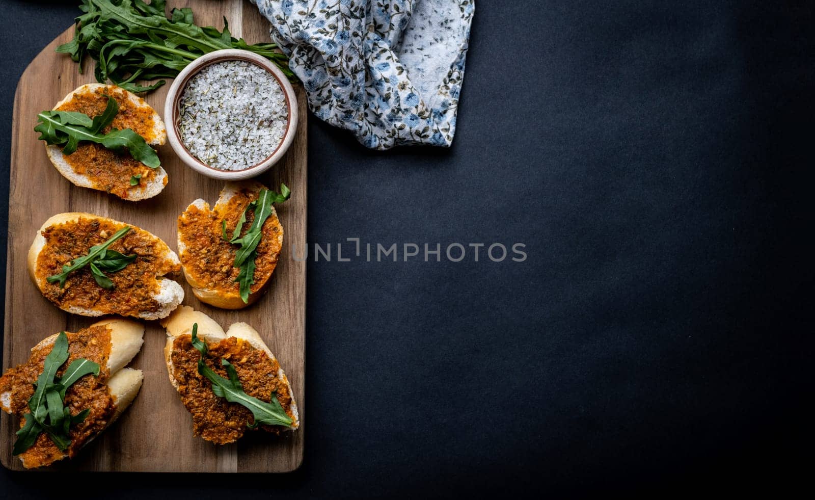 Mediterranean bruschettas with pesto and arugula served on wooden board with herbal salt and fabric on background with copy space. Traditional italian toasted bread with sauce and plants antipasti