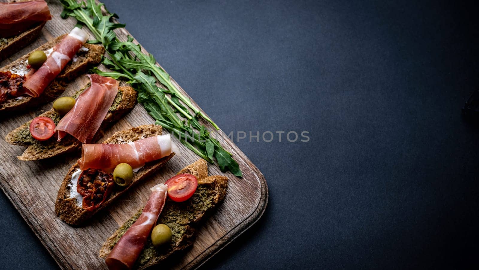 Bruschettas with jamon, olives, pesto, grilled cherry tomatoes served on wooden board with arugula on background with copy space. Mediterranean toasted bread with meat, cheese and vegetables