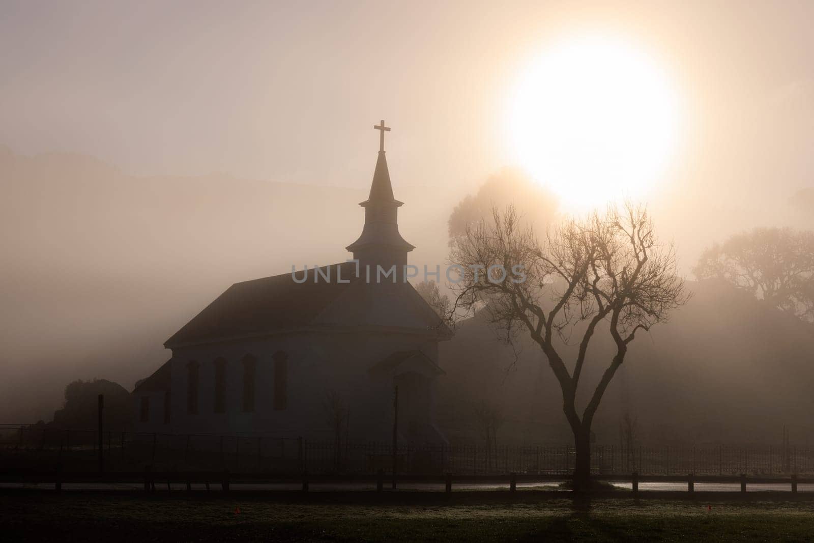 Brilliant bright sun silhouettes small church and tree in fog at dawn. High quality photo