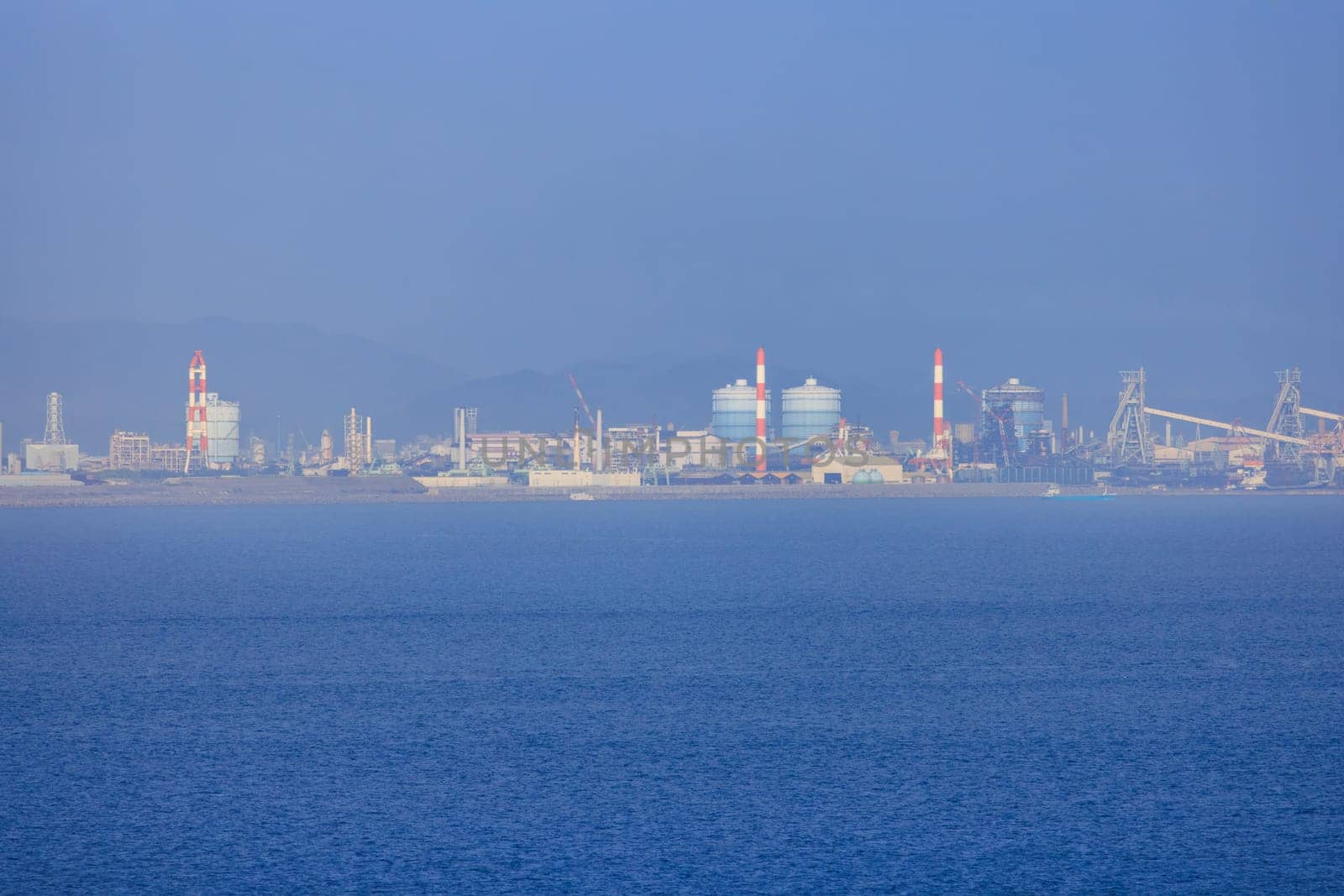 Red and white chimneys at industrial plant on coast in hazy atmosphere by Osaze