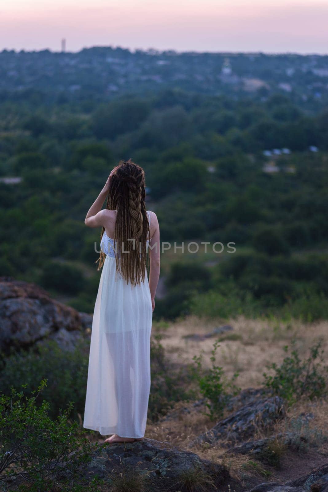 A beautiful girl in a white dress with dreadlocks is standing on a rock. Back view. Against the sunset sky. by Renisons