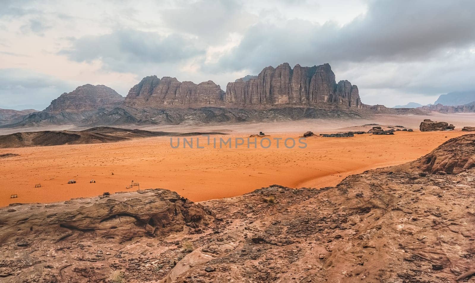 Rocky desert scenery in Wadi Rum desert during overcast morning, camp tents visible in distance.