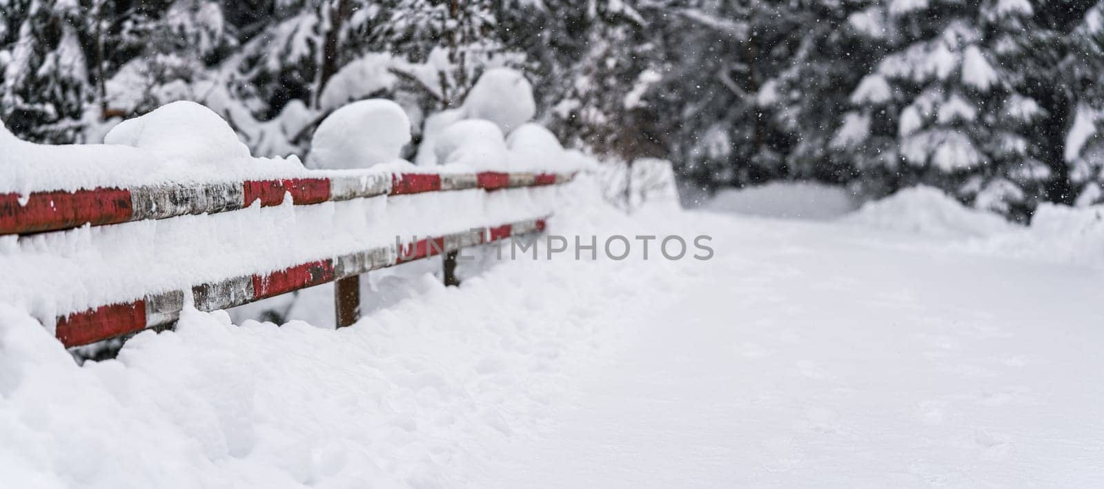 Red and white painted metal safety fence on side of snow covered forest road bridge - wide photo, space for text right side. Winter car driving background.