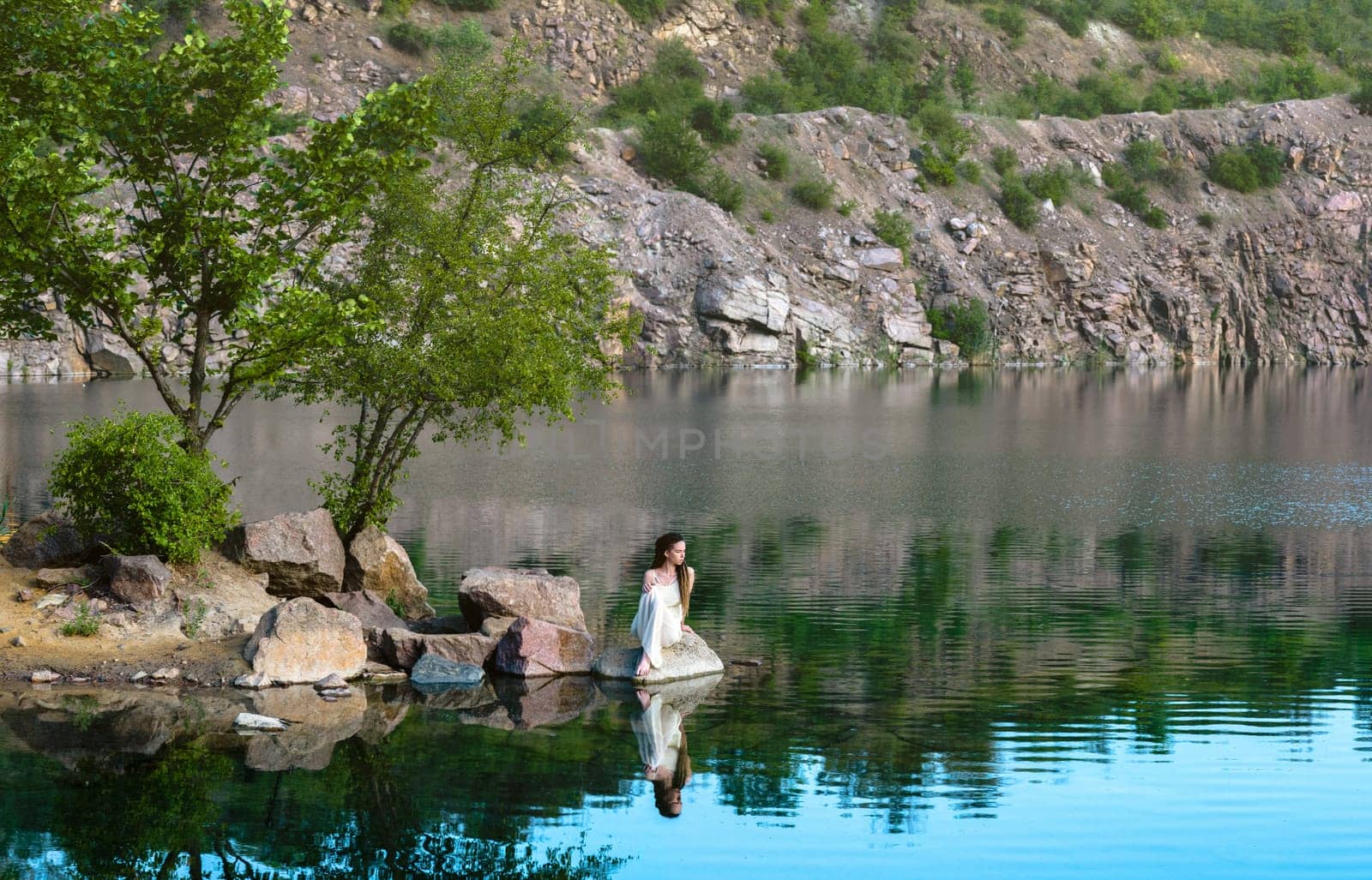 Beautiful girl sitting on a rock in a light dress. On the shore of the lake. by Renisons