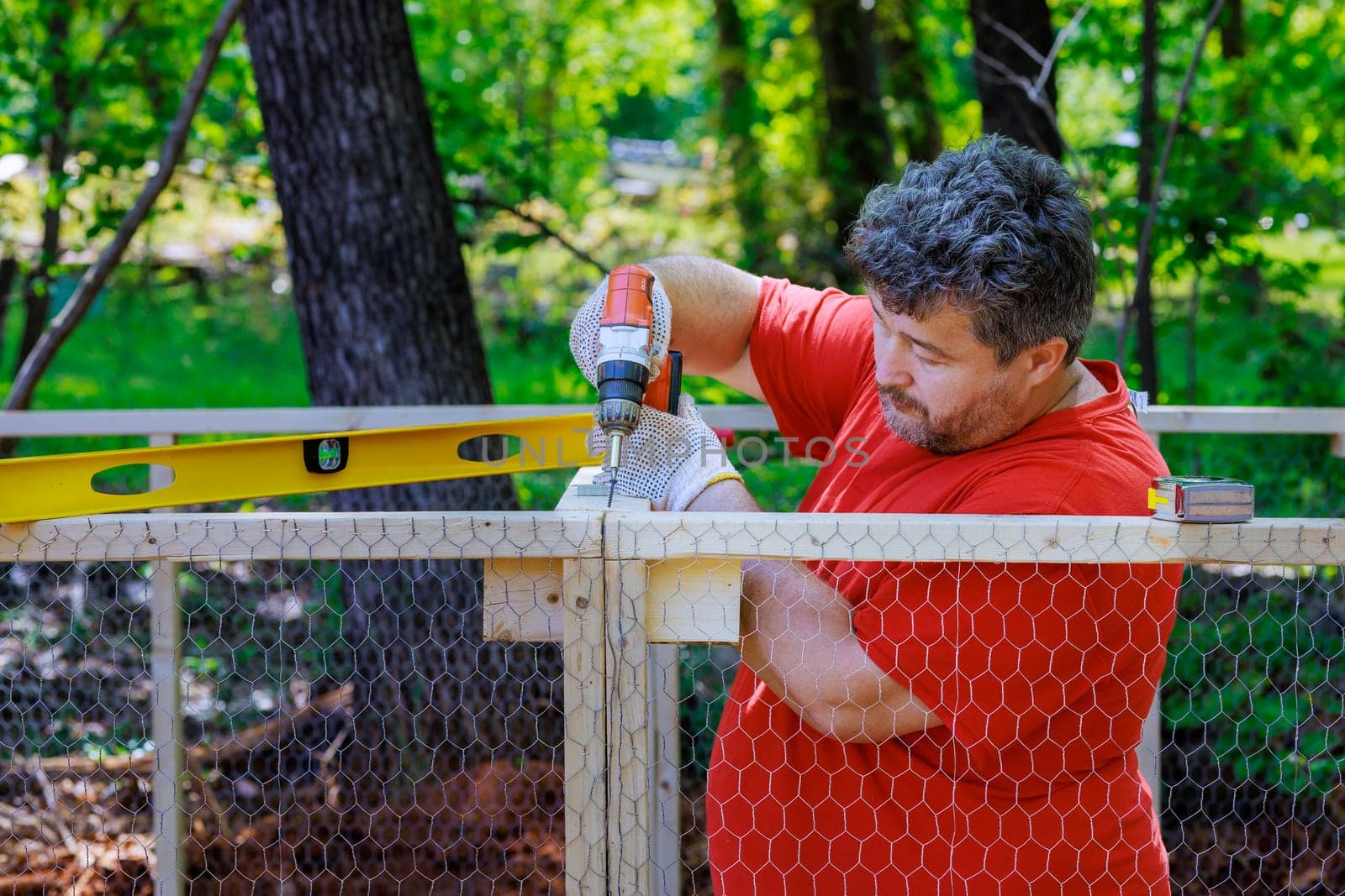 On a farm, a worker uses a screwdriver to install a metal grid in a wooden chicken coop that has been constructed from wood by ungvar