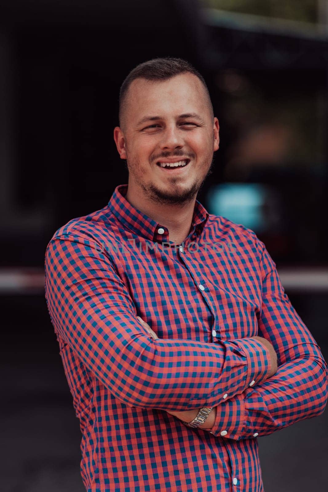 A successful young businessman in a shirt, with crossed arms, poses outdoors, confident expression on his face