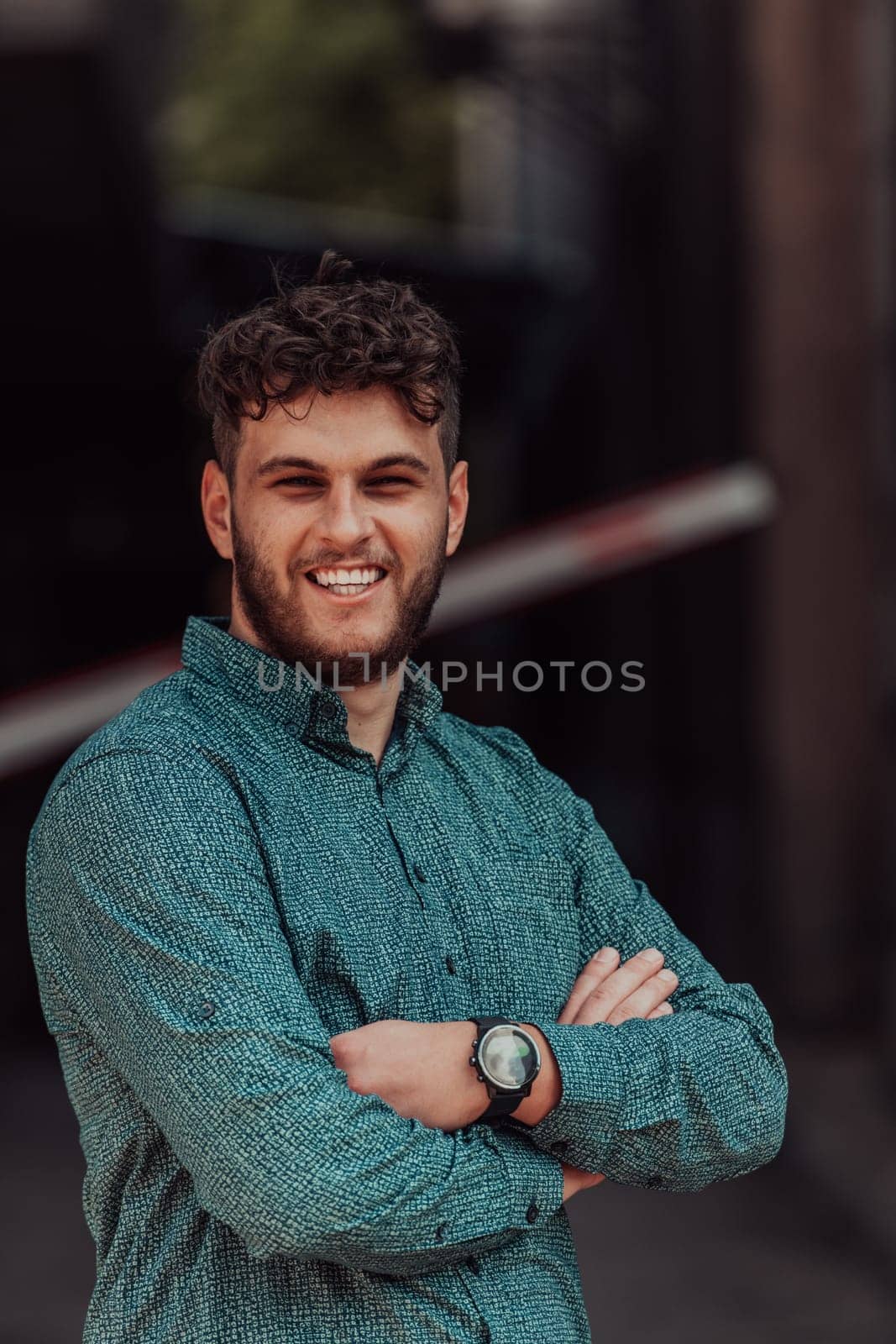 A successful young businessman in a shirt, with crossed arms, poses outdoors, confident expression on his face