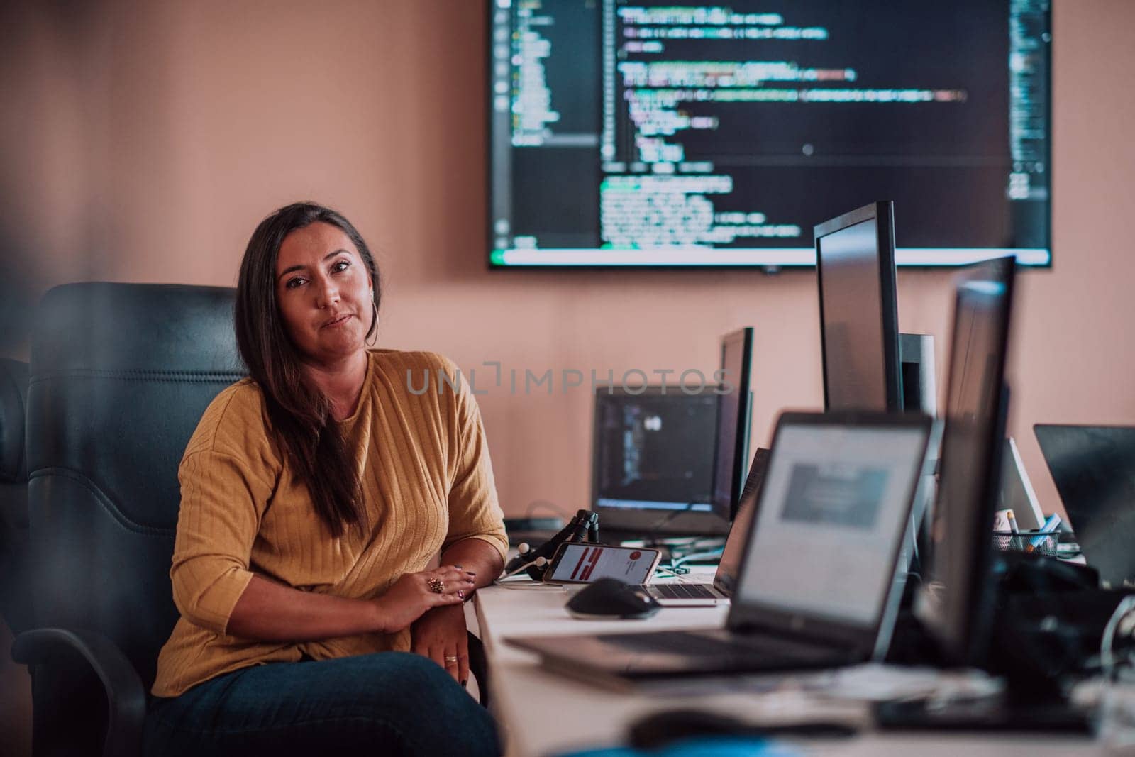 A businesswoman sitting in a programmer's office surrounded by computers, showing her expertise and dedication to technology. by dotshock