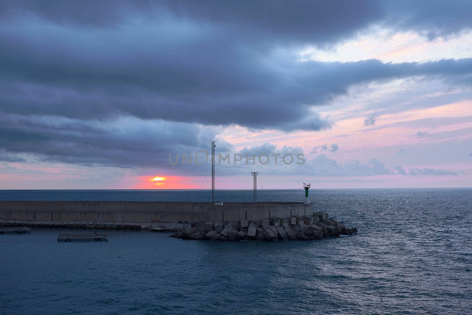 Sunrise from green lighthouse on maritime breakwater.Rocks, jetty, blue and cold tones, orange sunshine