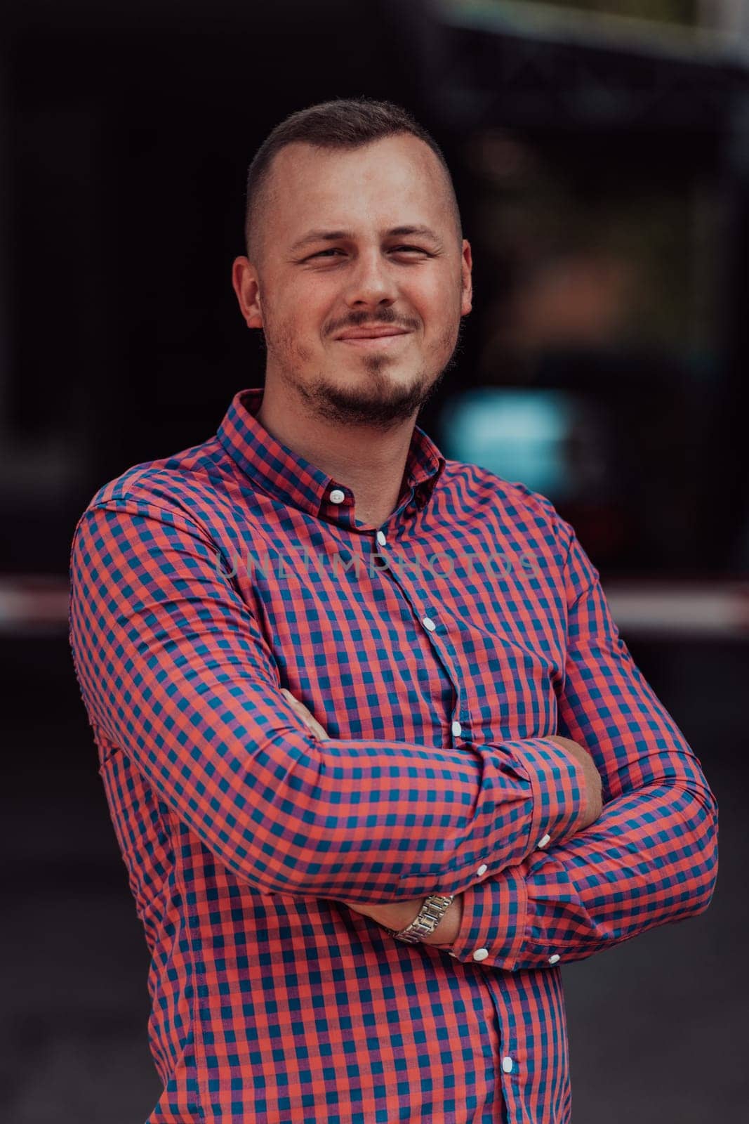 A successful young businessman in a shirt, with crossed arms, poses outdoors, confident expression on his face