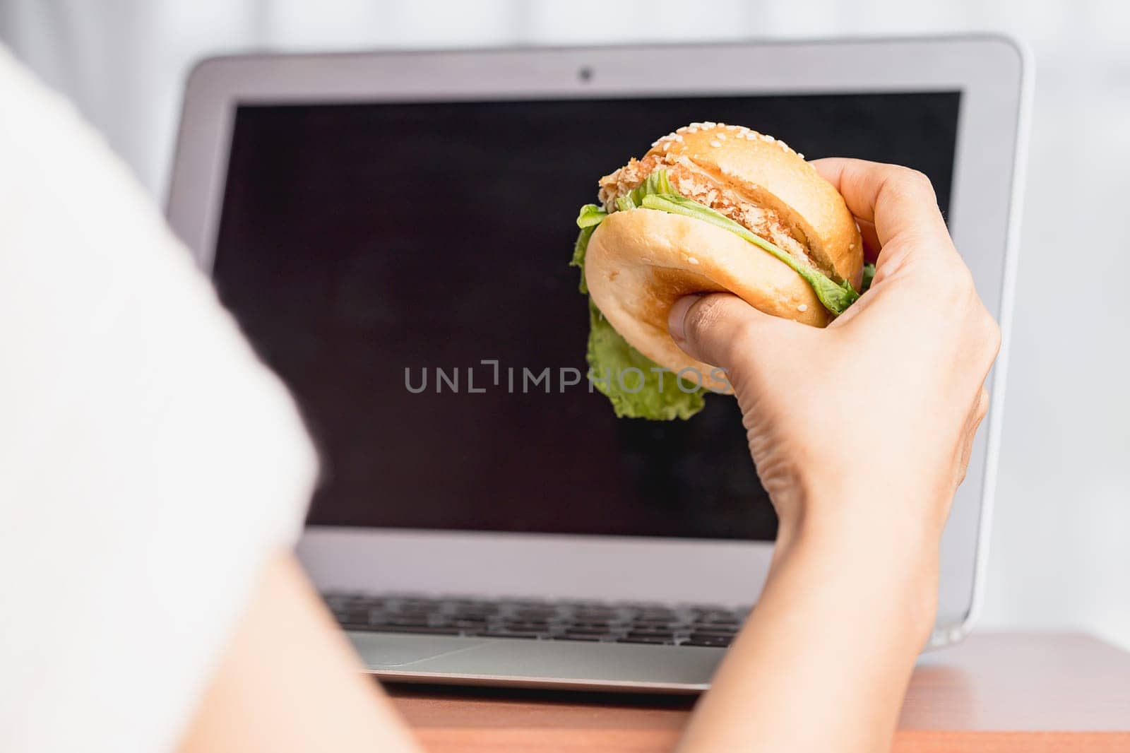 Hand holding a delicious hamburger and using laptop computer for eating while working concept.