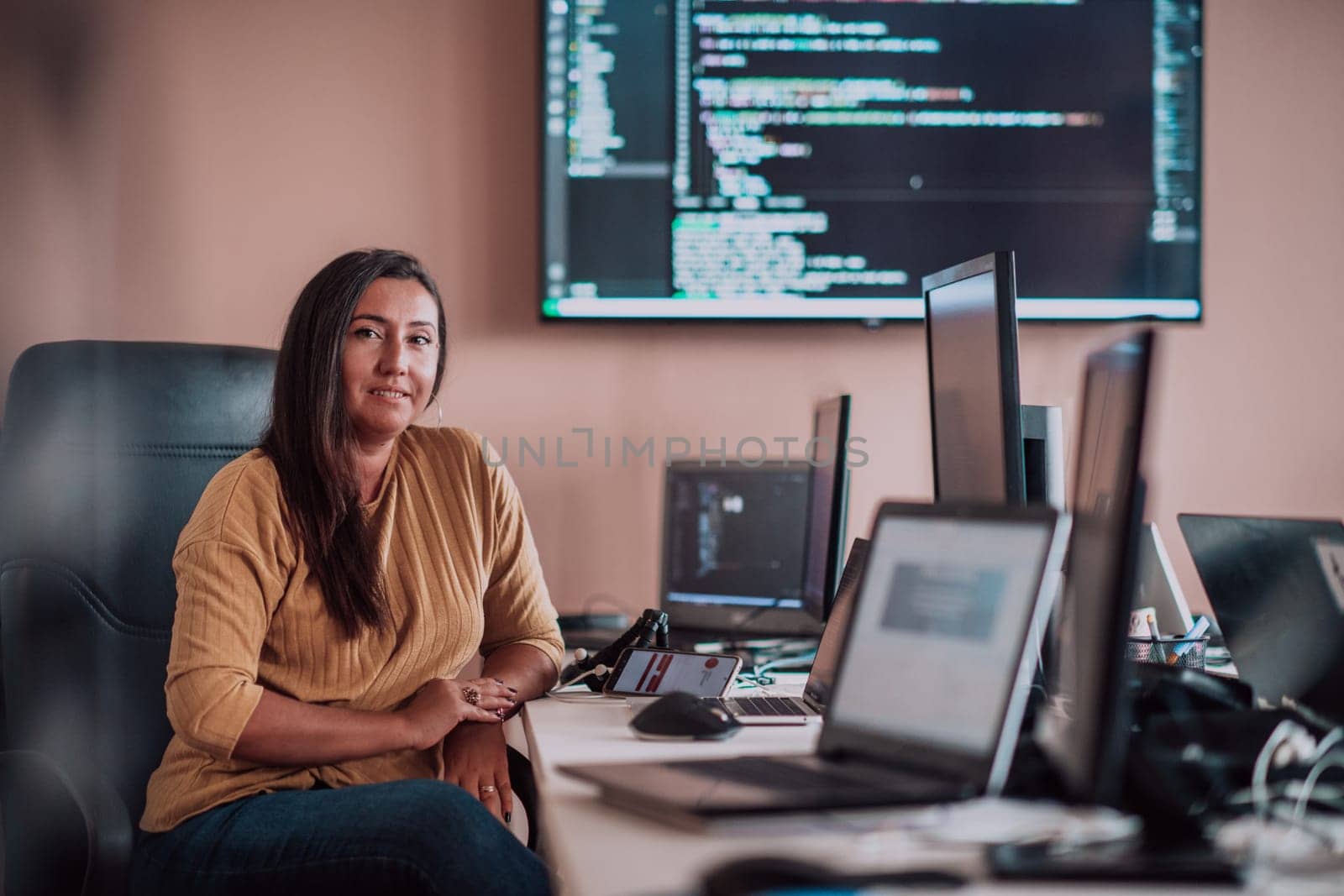 A businesswoman sitting in a programmer's office surrounded by computers, showing her expertise and dedication to technology