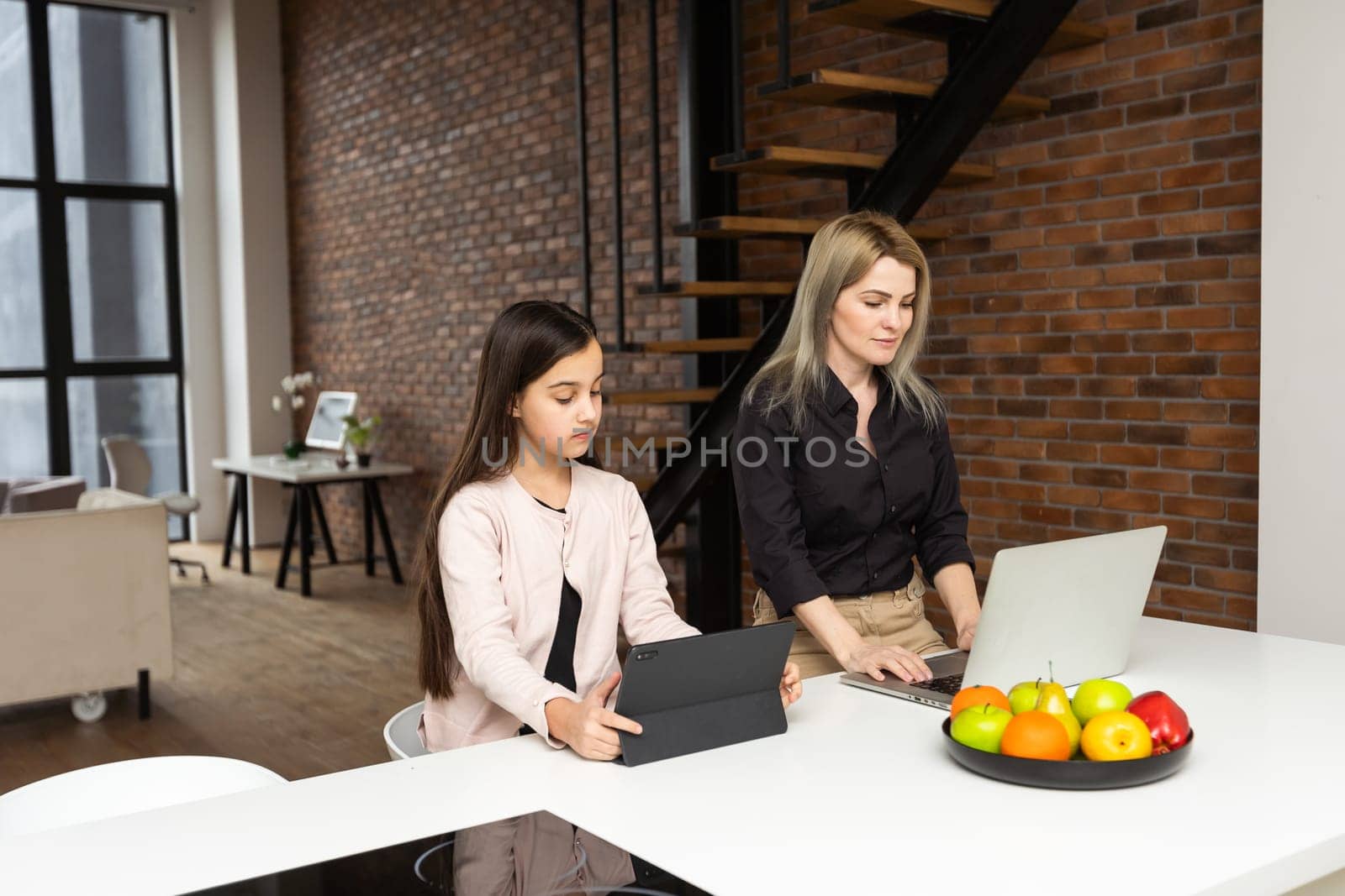 Portrait of mother and daughter sitting in living room. Cute girl holding in her hand a digital tablet while her mom using laptop and looking at something together