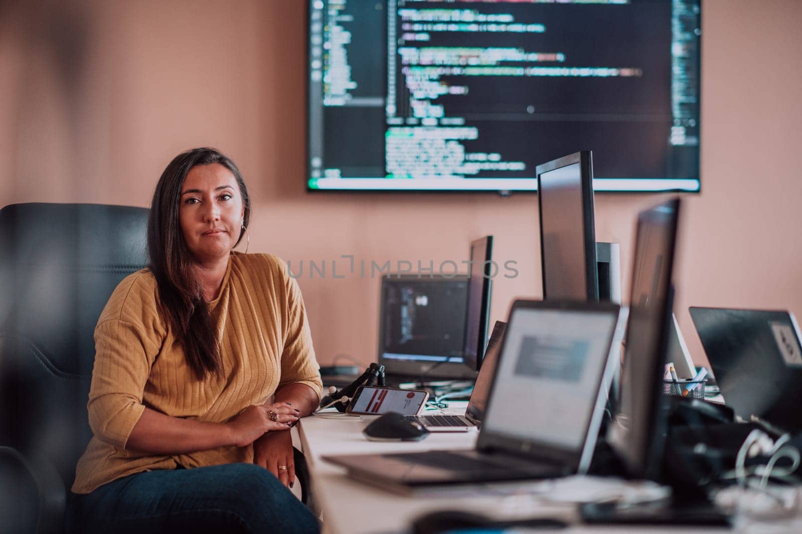 A businesswoman sitting in a programmer's office surrounded by computers, showing her expertise and dedication to technology