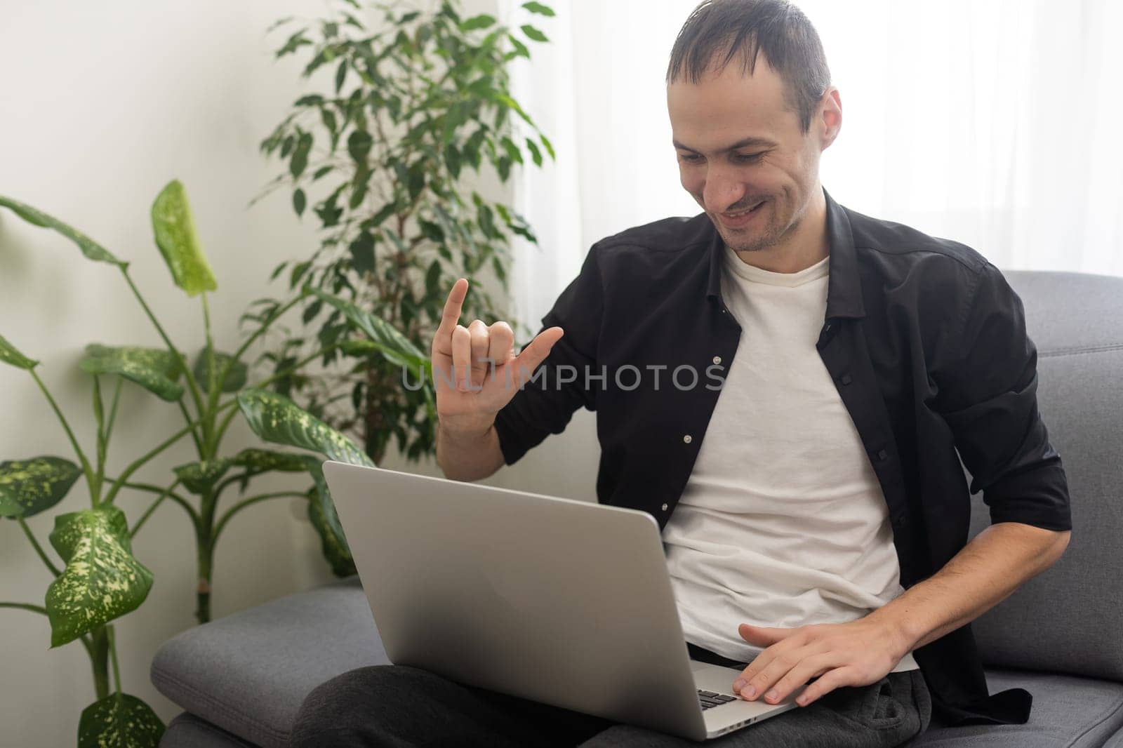 Deaf or hard hearing happy smiling young caucasian man uses sign language while video call using laptop while sitting on the couch at home