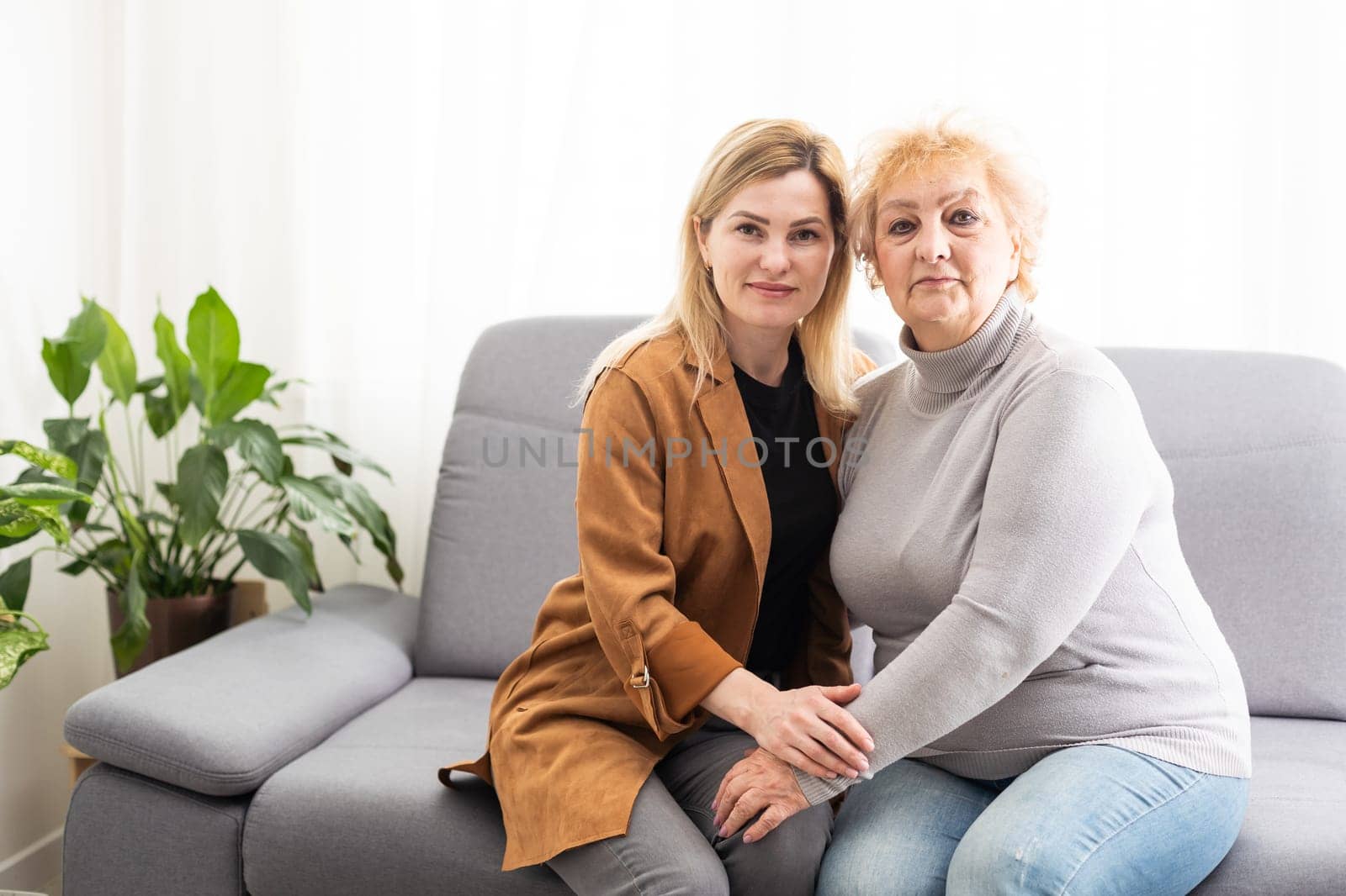 Close up image aged mother adult daughter sitting on couch indoors smiling looking posing for camera hugging feels happy spend time together, concept of love relative people, multi-generational family by Andelov13
