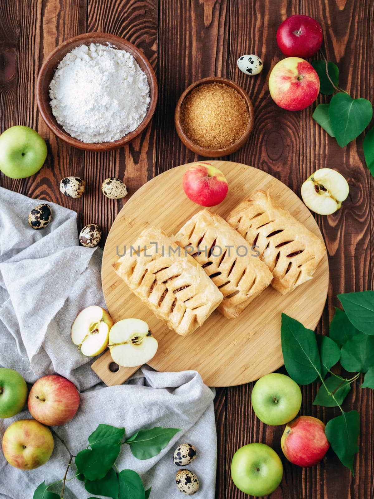 Autumn baking concept. Top view food photography. Baked puff pastry crispy apple puff with ingredients on a rustic wooden table. Puff pastry pies with apples, brown sugar, eggs, flour and towel.