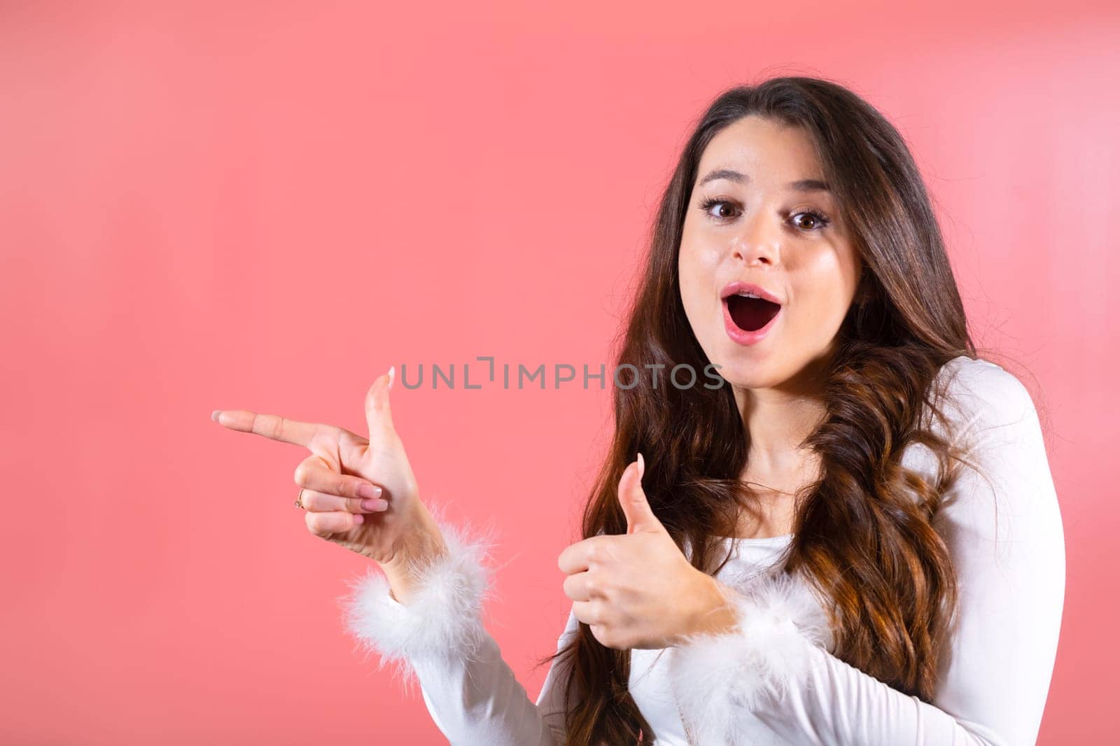 Portrait of an exciting young woman gracefully gesturing with her fingers, showing on the copy space on the pink background