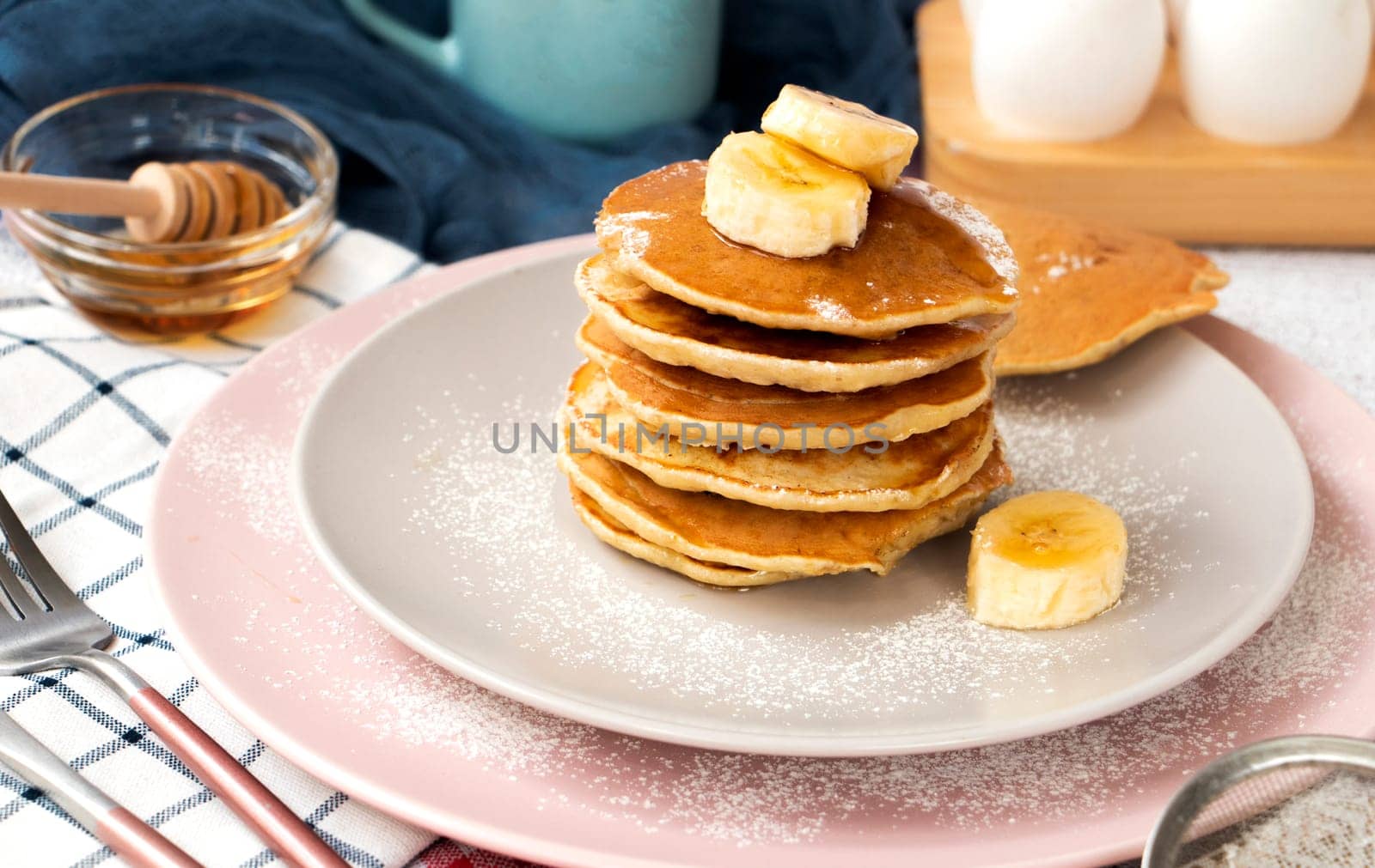 homemade banana pancakes on plate sprinkled powder and honey. Pancakes on the kitchen table against the background of eggs and cups with coffee. Pancake slide with honey topping and fresh bananas