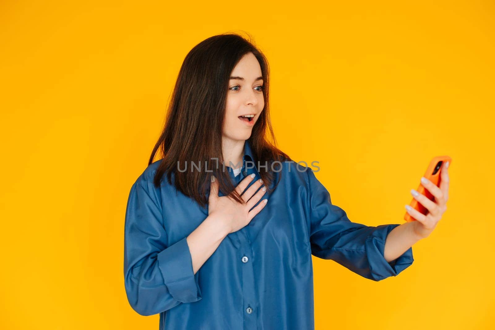 Digital Surprise: Captivating Young Woman in Stylish Shirt, Reading Astonishing News on her Phone - Vibrant Expression Isolated on Yellow Background