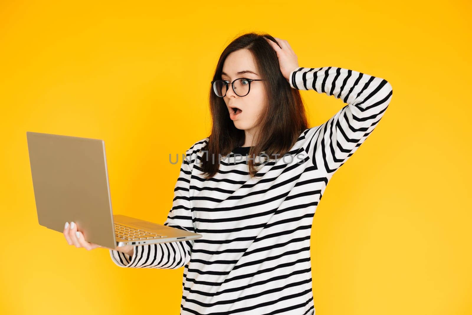 Picture of a cute impressionable lady in a striped T-shirt with glasses, reading the news or writing on a modern laptop device, hand head isolated yellow background color by ViShark