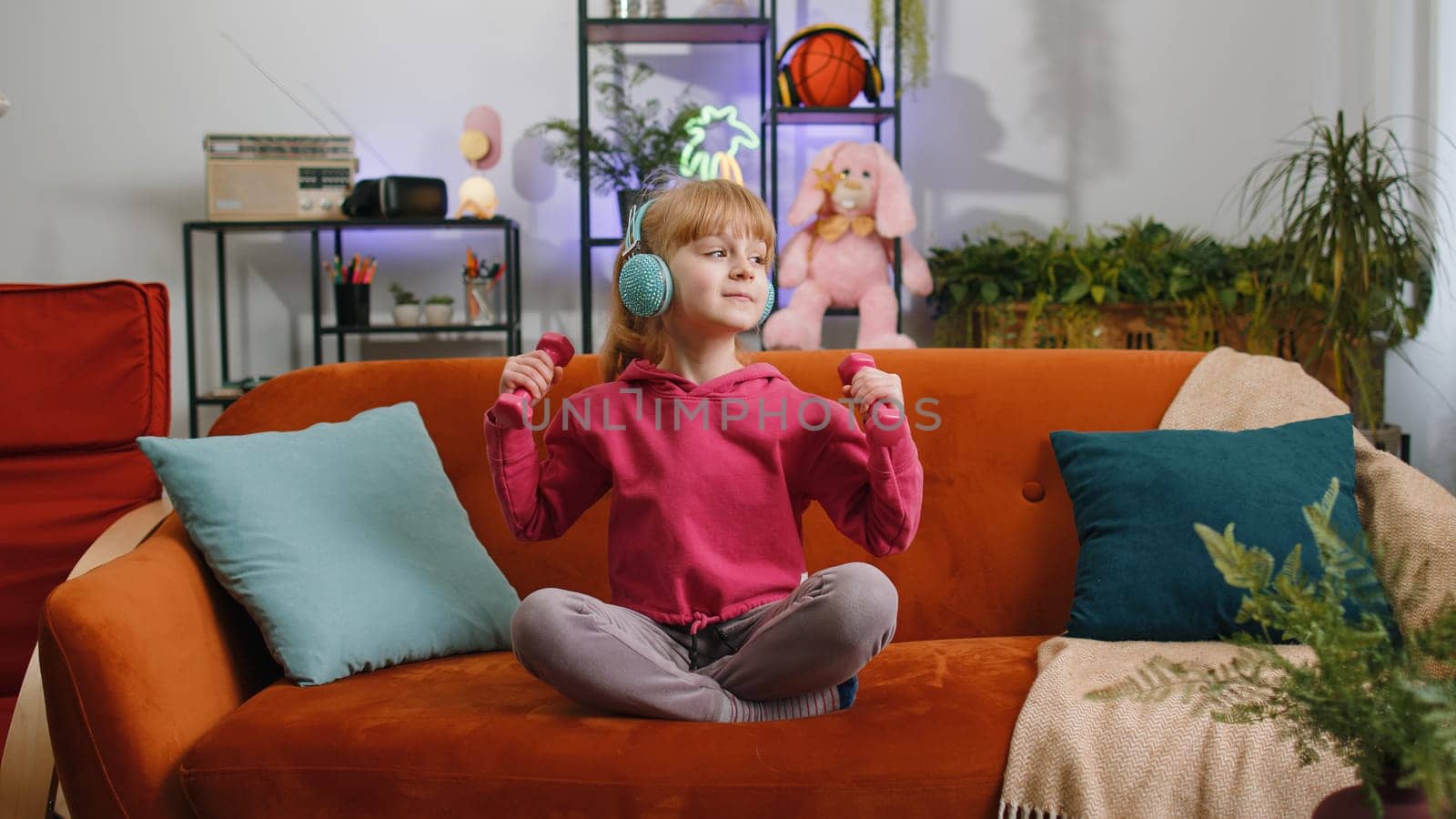 Young blonde child kid girl lifting dumbbells while doing exercises, enjoying training. Female teen toddler wearing headphones listening music at home in living room sits on sofa. Sport and fitness