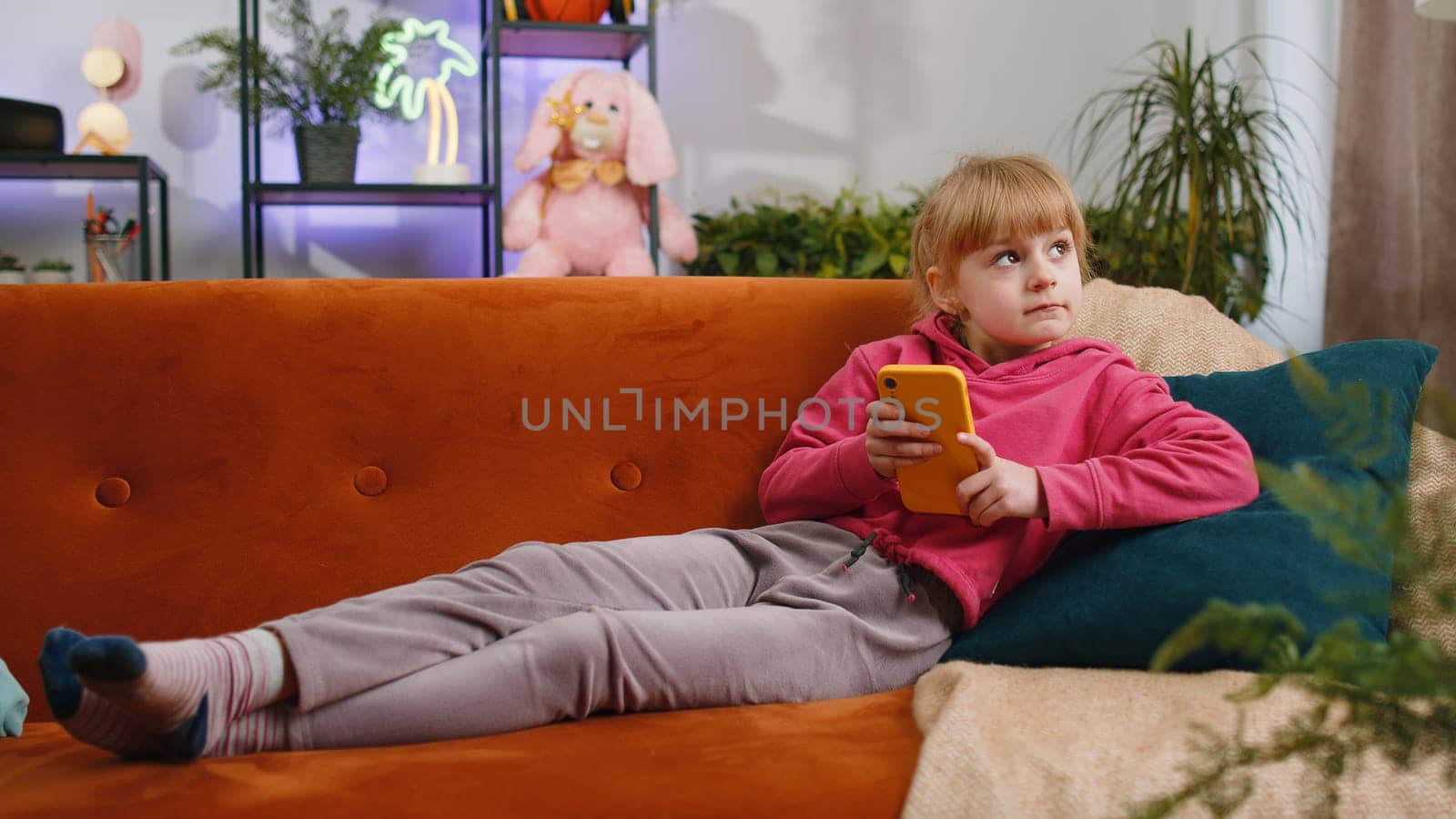 Young child kid girl texting share messages content on smartphone social media applications online, watching relax movie. Female teen toddler uses mobile phone at home in living room lying on sofa