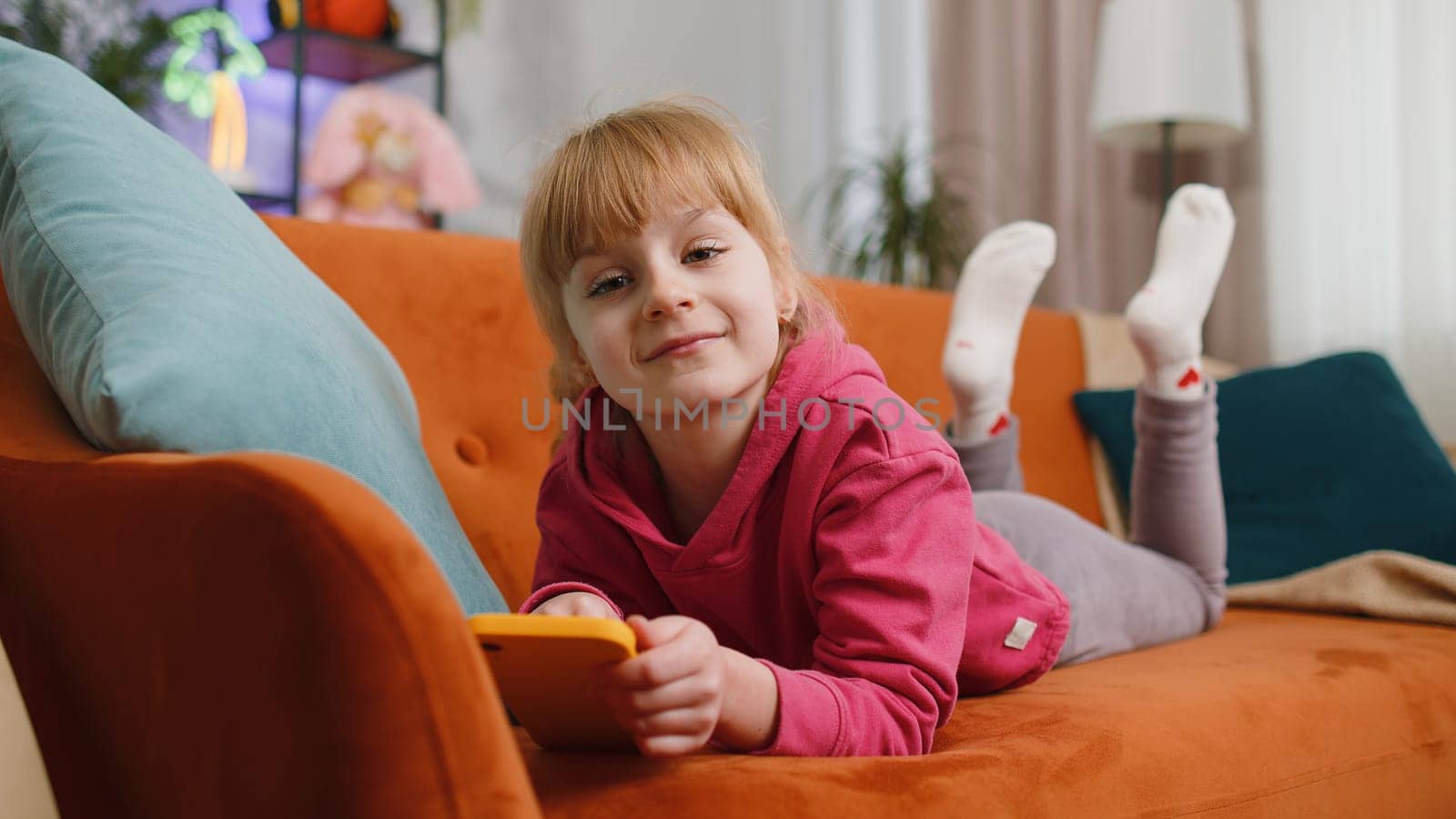 Young child kid girl texting messages on smartphone social media applications, watching relax movie, online shopping, play game. Female teen toddler uses mobile phone at home in room lying on sofa