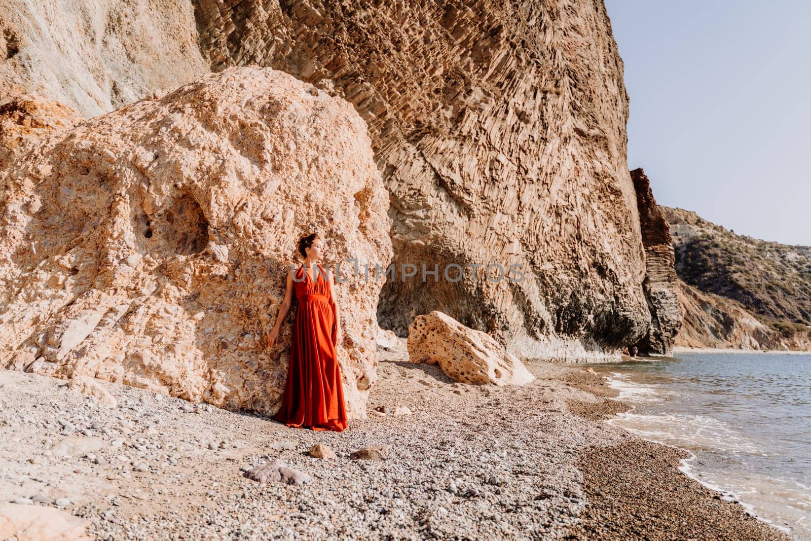 Woman red dress sea. Woman in a long red dress posing on a beach with rocks on sunny day. Girl on the nature on blue sky background