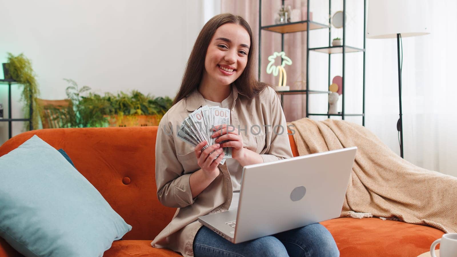 Planning budget. Rich happy young woman counting money cash use laptop computer calculate domestic bills sitting on sofa. Girl satisfied of income earnings, saves money for planned vacation, gifts