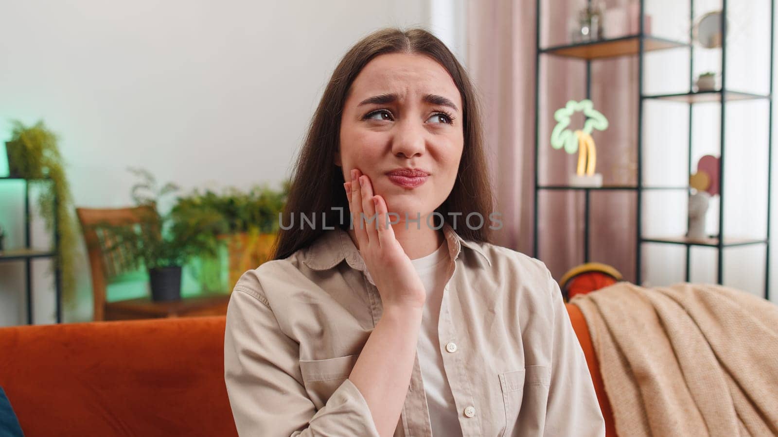 Dental problems. Portrait of young woman touching cheek, closing eyes with expression of terrible suffer from painful toothache, sensitive teeth, cavities. Girl sitting on sofa at home room apartment