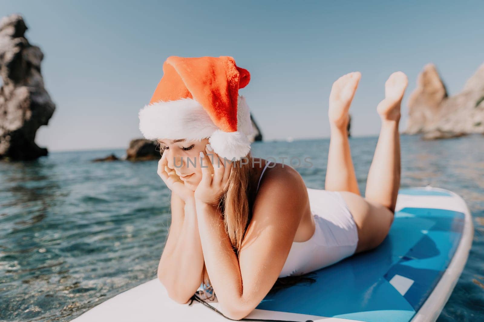 Woman sea sup. Close up portrait of happy young caucasian woman with long hair in Santa hat looking at camera and smiling. Cute woman portrait in a white bikini posing on sup board in the sea by panophotograph