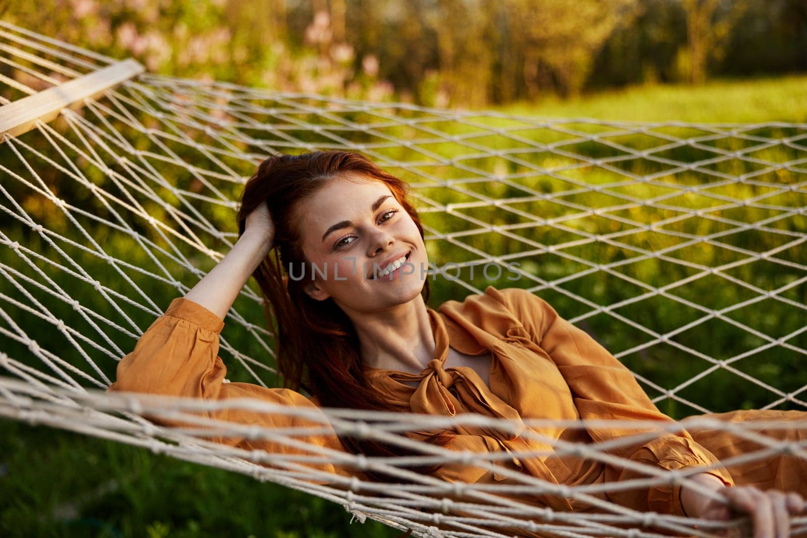 a happy woman is resting in a mesh hammock, resting her head on her hand, smiling happily looking at the camera, enjoying a sunny day. High quality photo