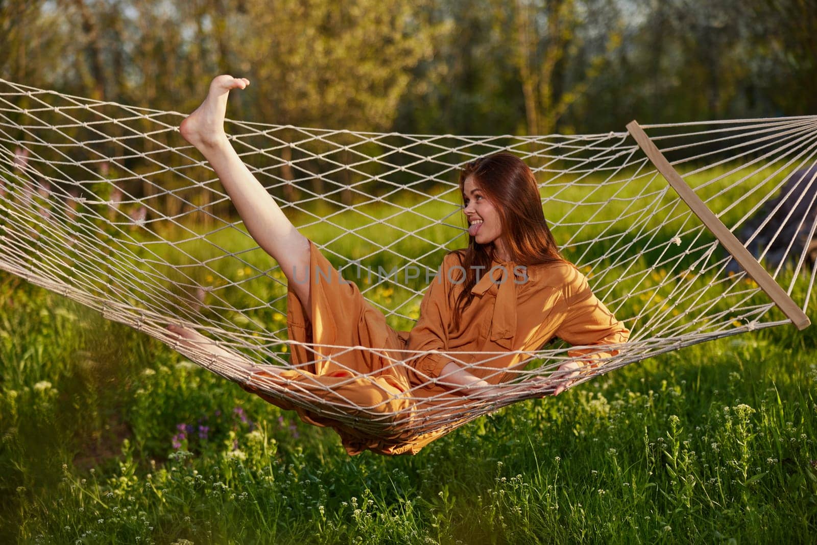 horizontal photo of a beautiful, red-haired woman lying in a hammock enjoying a rest in a long orange dress, on a warm summer day, funny lifting her leg. High quality photo