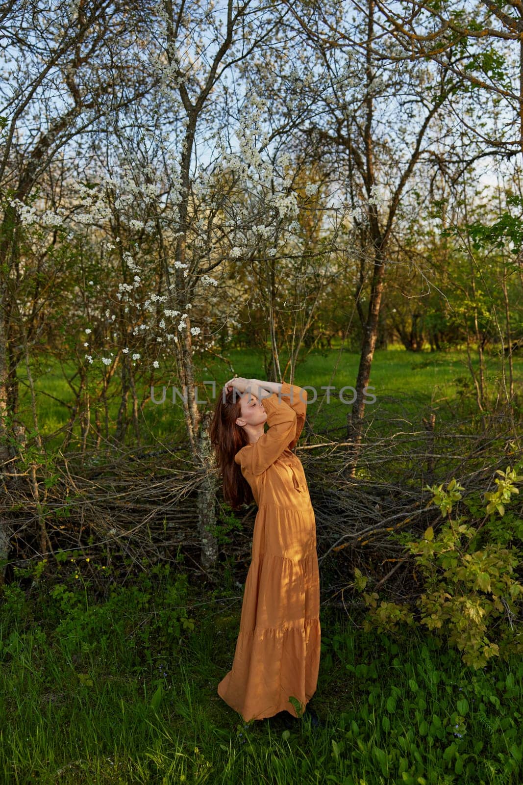an elegant, sophisticated woman poses relaxed standing near a wicker fence at the dacha in a long orange dress touching her head with her hands by Vichizh