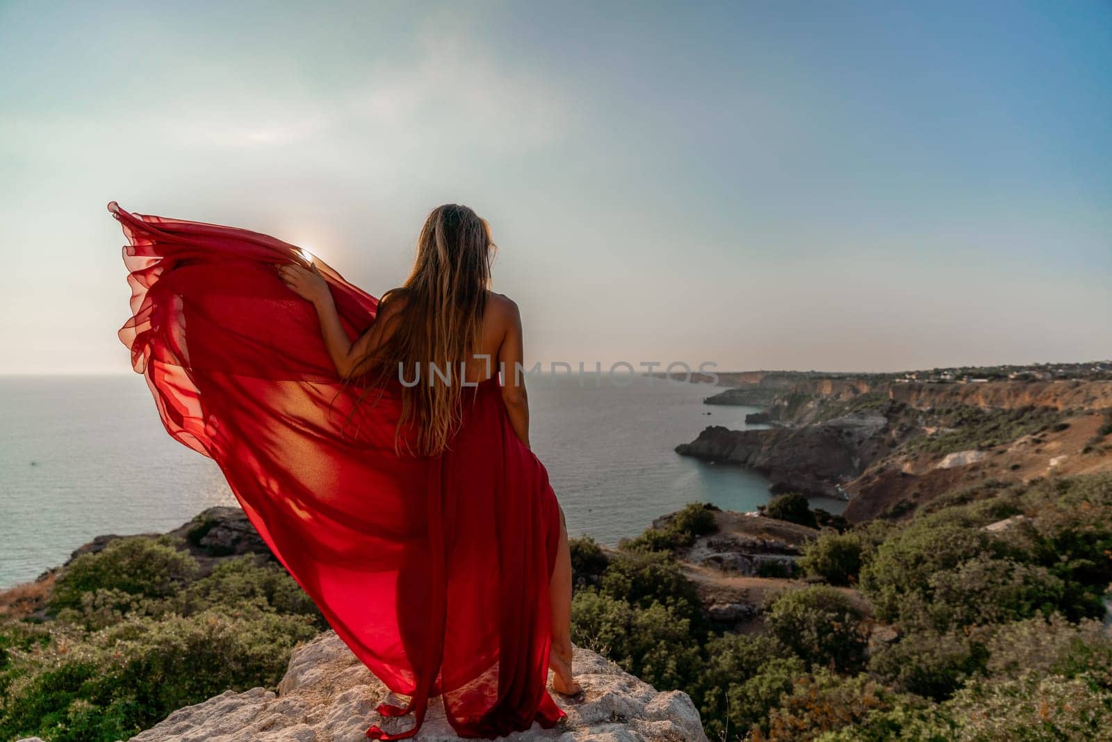 Woman sunset sea red dress, back view a happy beautiful sensual woman in a red long dress posing on a rock high above the sea on sunset. by Matiunina