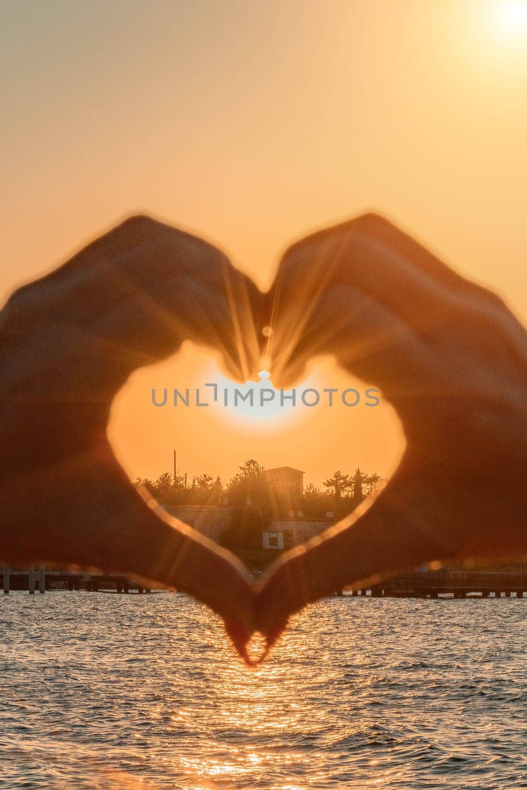 Silhouette against the sunset - hands folded in the shape of a heart. Sun with rays between the palms. Concept for Valentine's Day, declaration of love. Minimalism, space for text, warm colors. by Matiunina