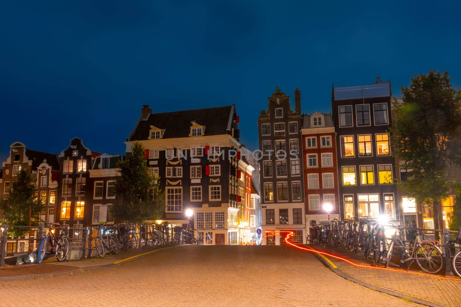 Netherlands. Summer night in Amsterdam. Bicycles are parked at the fence of the bridge over the canal and typical Dutch buildings on the quay