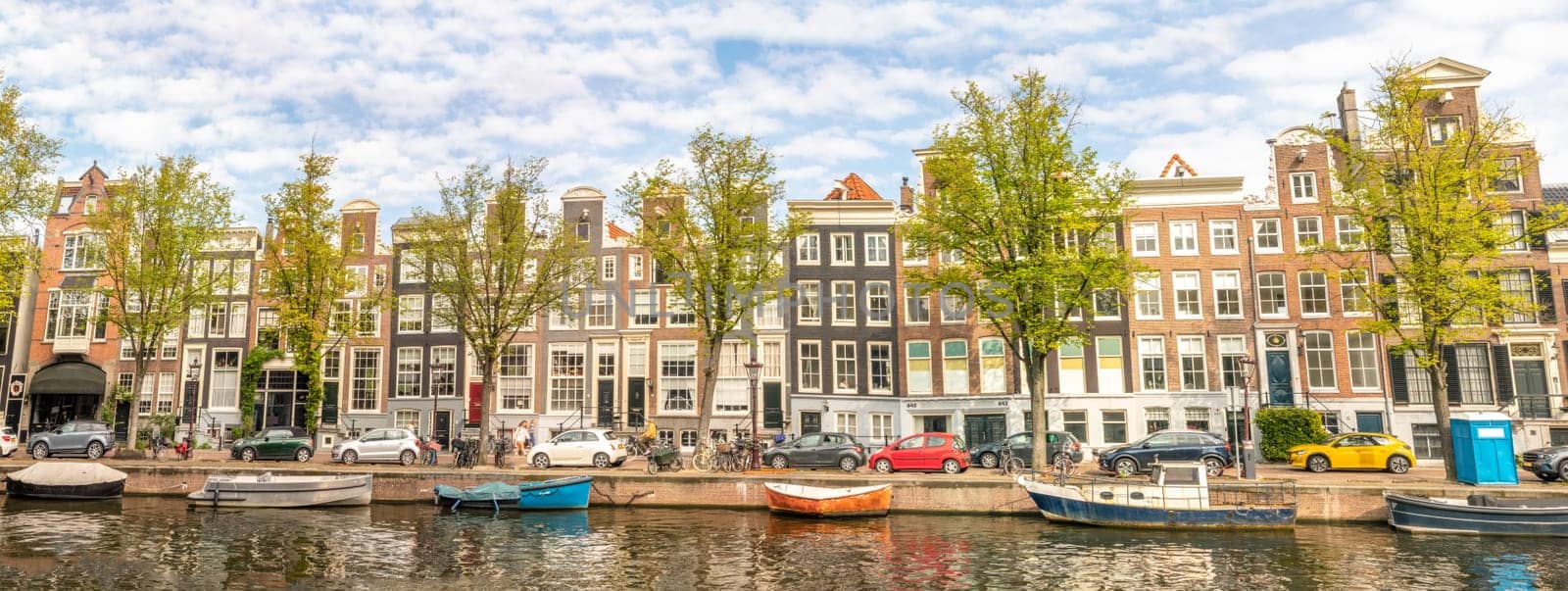 Netherlands. Sunny summer day in Amsterdam. Panorama of the canal quay. Boats and cars are parked in front of the facades of typical Dutch buildings