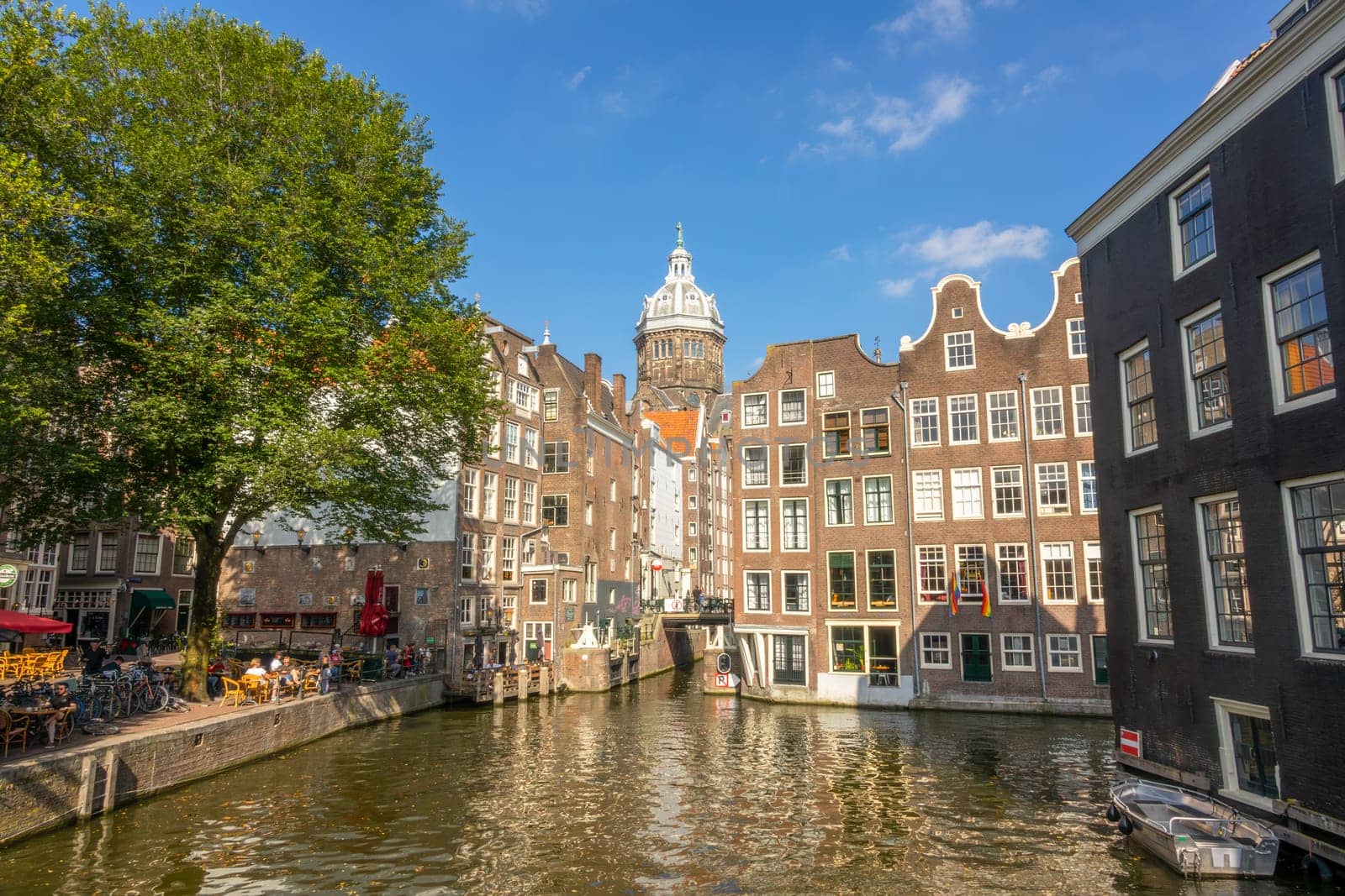 Netherlands. Sunny summer day in Amsterdam. Typical Dutch houses near the canal water. Small street cafe in the shade of a big green tree