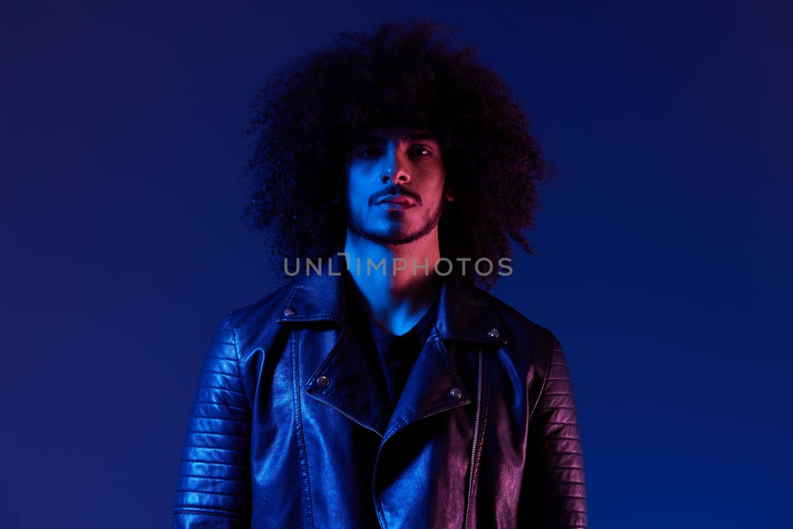 Portrait of fashion man with curly hair on blue background multinational, colored light, black leather jacket trend, modern concept. High quality photo