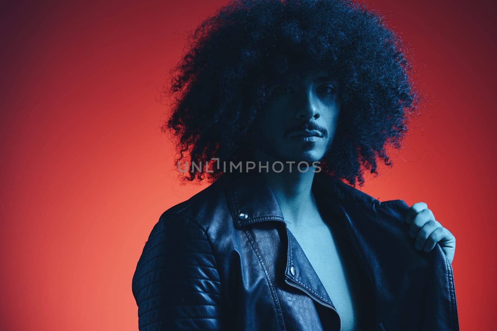 Portrait of fashion man with curly hair on red background multinational, colored light, black leather jacket trend, modern concept. High quality photo