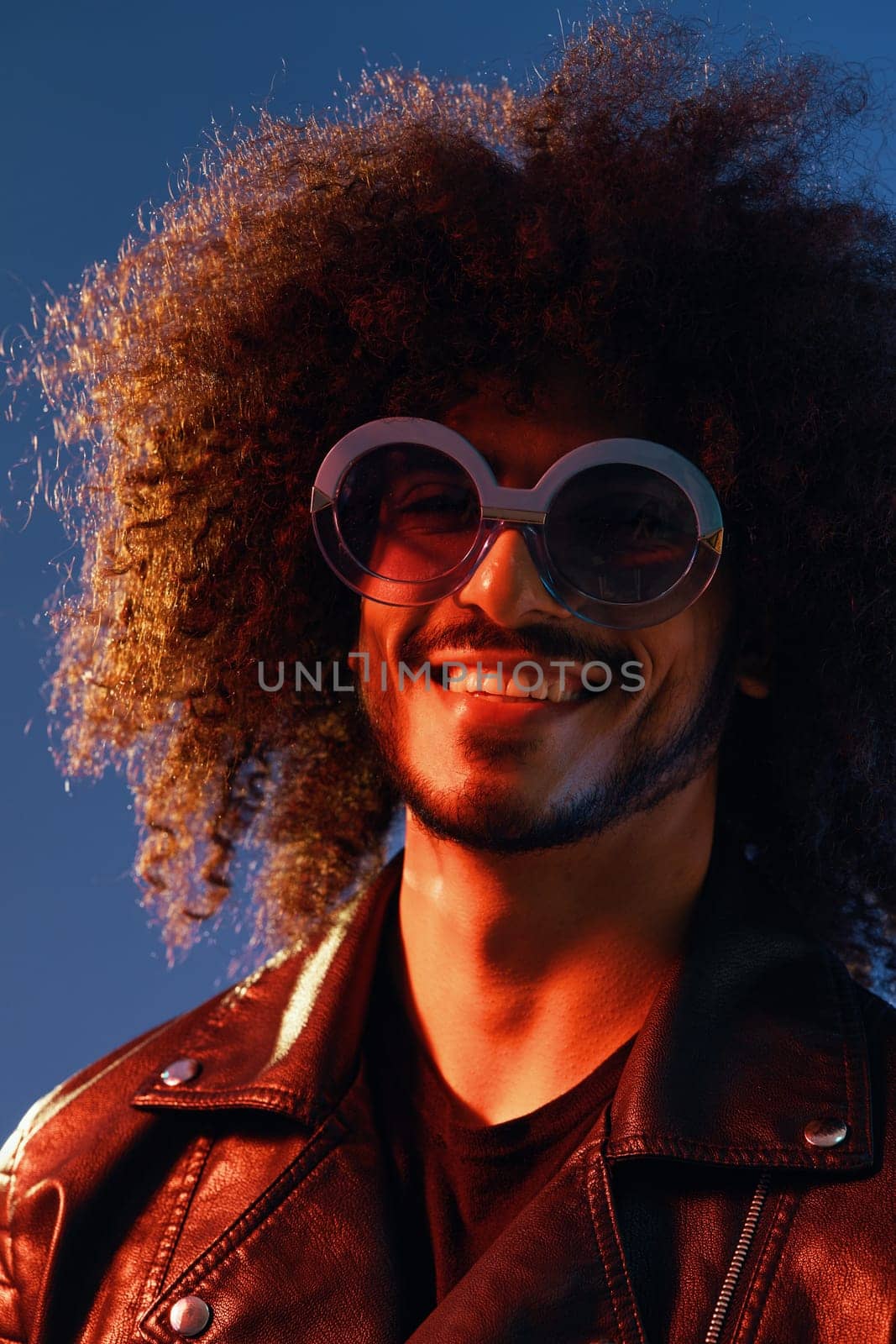 Portrait of a stylish man with curly hair with glasses smile on blue background multinational, color light, black leather jacket trend, modern concept. High quality photo